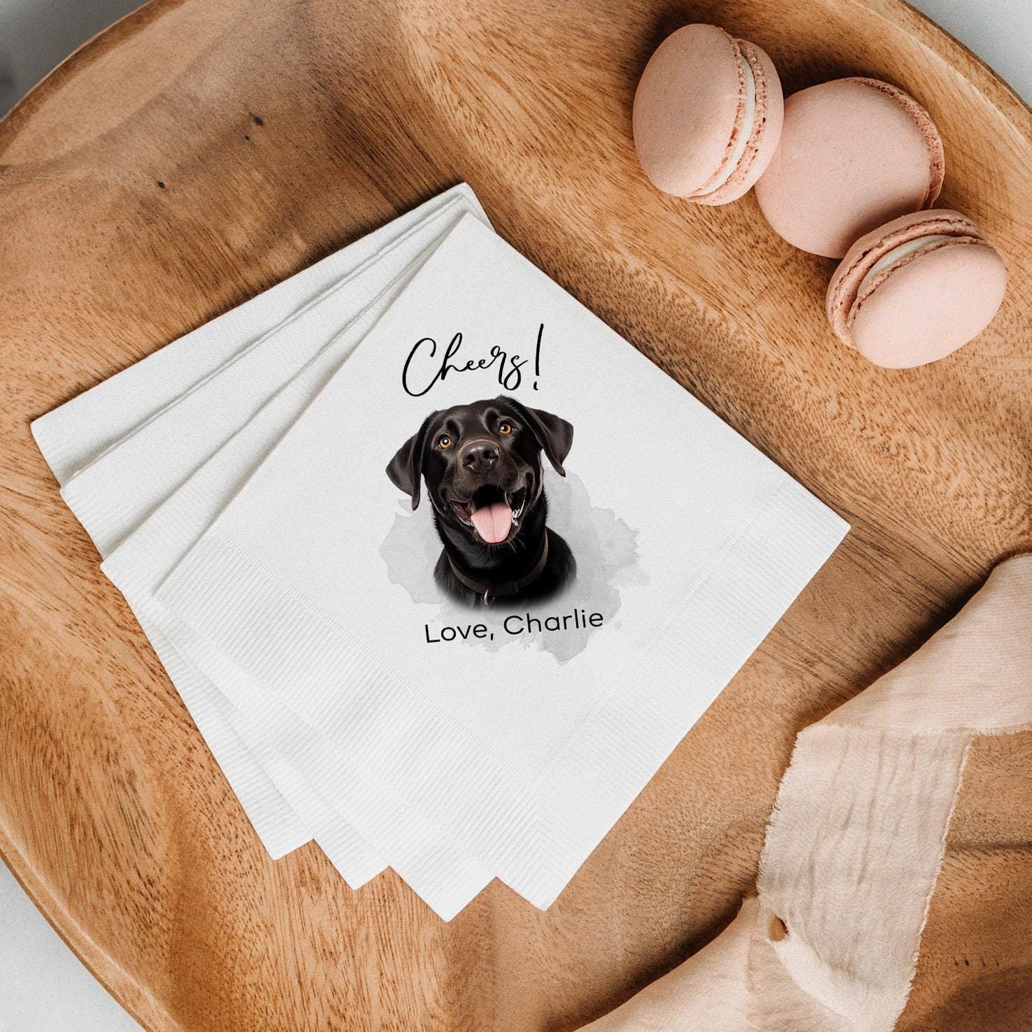 White cocktail napkins featuring a watercolor-style black Labrador's face with text "Cheers! Love, Charlie," placed on a wooden tray alongside blush-colored macarons and soft beige fabric.
