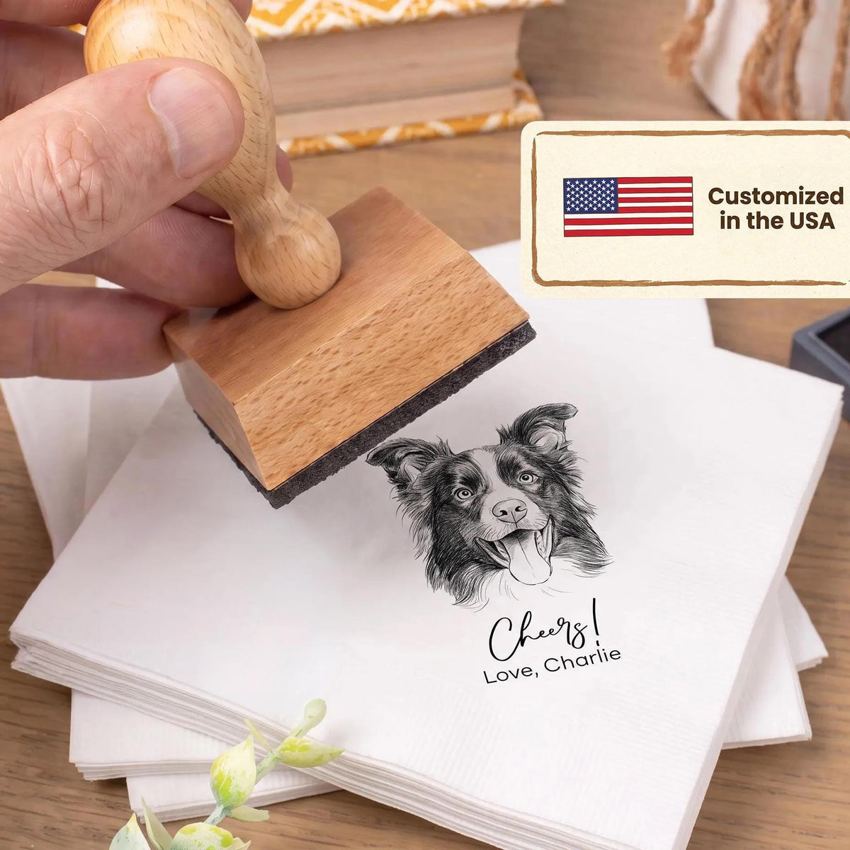A hand is holding a wooden stamp with an image of a dog's face, and the stamp is being used to print a personalized message on a stack of white napkins.