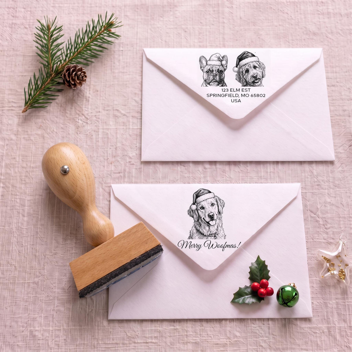 two white envelopes with black illustrations of dogs wearing Santa hats, along with a wooden stamp and holiday decorations such as pine branches, a pine cone, and holly leaves.