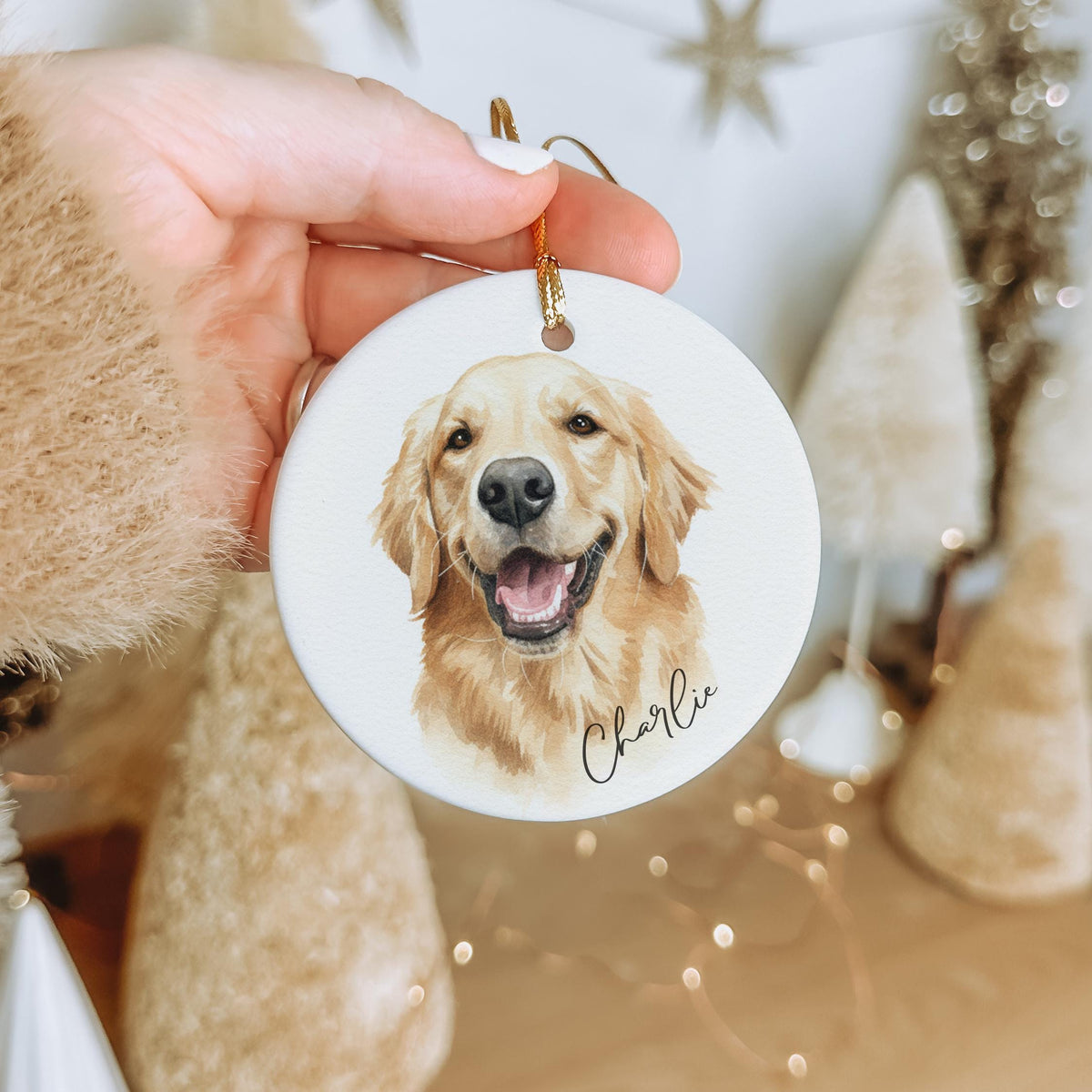 Personalized round ceramic Christmas ornament featuring custom watercolor portrait of a golden retriever dog and name Charlie, hand holding ornament with cozy neutral holiday background and festive decor