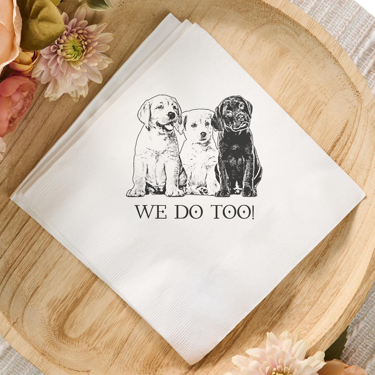 Custom white cocktail napkin featuring a black-and-white illustration of three dogs and the text 'WE DO TOO!' displayed on a wooden tray with pastel flowers, perfect for dog-themed weddings or events.