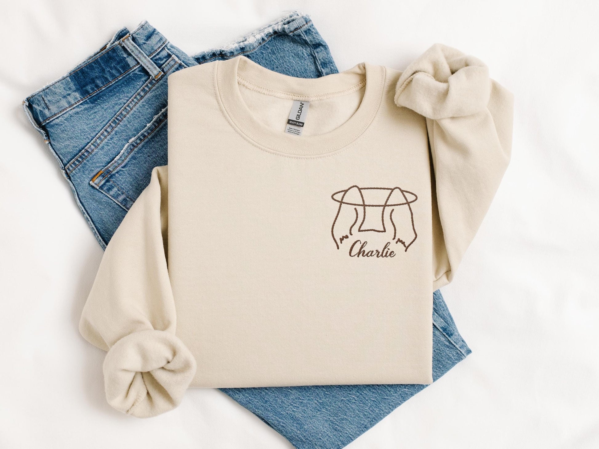 Beige sweatshirt with rolled sleeves, featuring a minimalist embroidered design of hands holding a pet&#39;s ears and the name &#39;Charlie,&#39; paired with blue denim jeans on a white background.