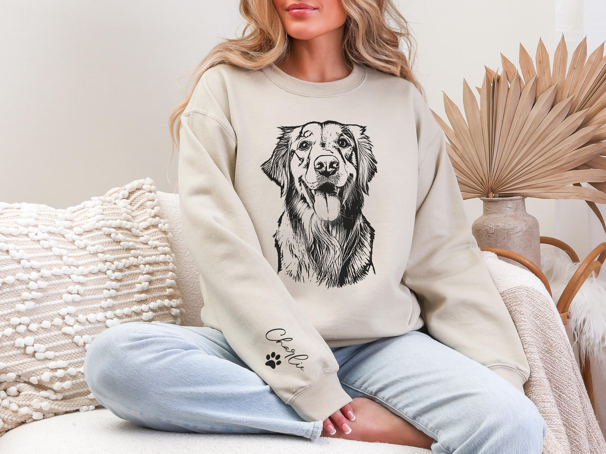 Woman wearing a beige custom pet sweatshirt featuring a detailed black line drawing of a dog’s face on the front, with personalized name “Charlie” and paw print on the sleeve, cozy home background with neutral decor.